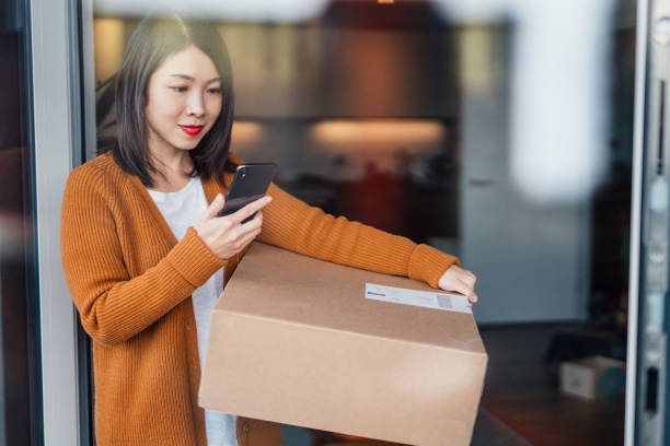 Young beautiful Asian woman holding a cardboard box, tracking parcel delivery using mobile app on smart phone. Home delivery service makes life easier. Online shopping concept.